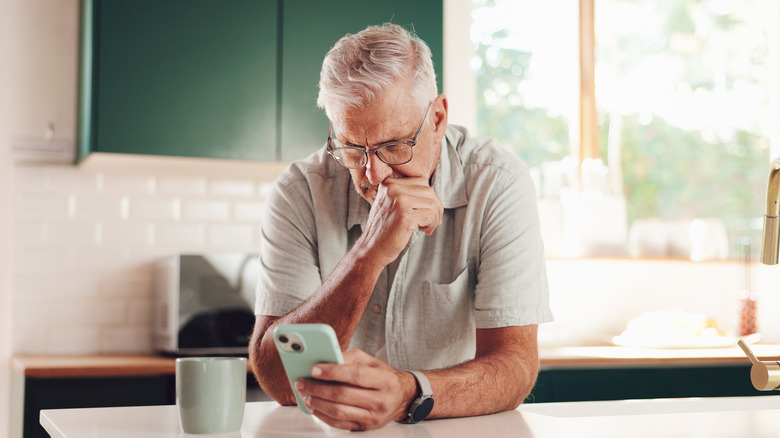 A senior mans looks worriedly at a phone, in a brightly lit kitchen