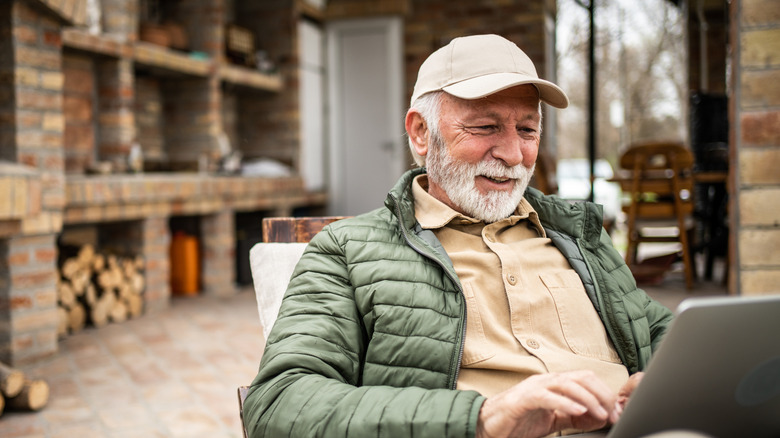 An older man smiles while working on his laptop on a rustic porch