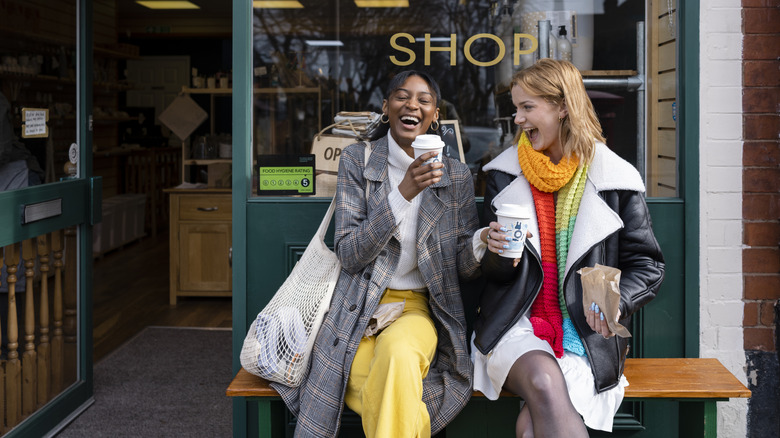 Two young women laughing together on a bench outside of a store