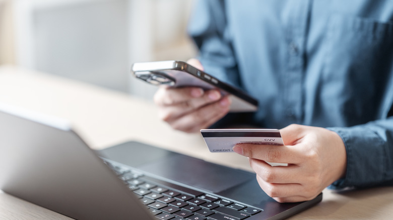hands holding a phone and card near laptop