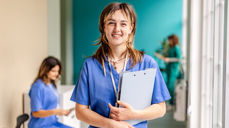 A young female smiling, wearing medical scrubs, and holding a file with other medical staff in the background.