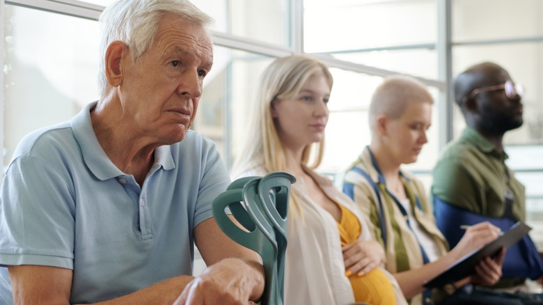 Old man sitting in a hospital queue with a crutch in one hand