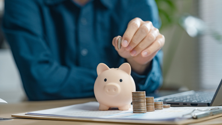 person in blue shirt putting quarters in piggy bank next to laptop