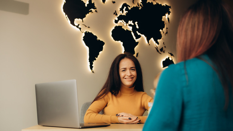 A smiling Gen Z woman sitting behind a desk in front of a client at a travel agency.