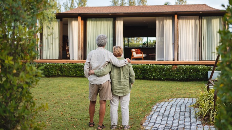 Older couple standing arm-in-arm on a lawn, looking toward a modern single-story home with large windows and a curved stone walkway
