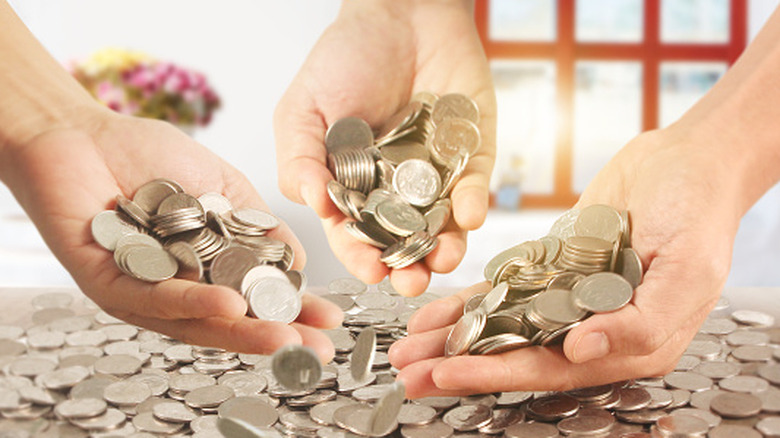 A closeup of three hands holding coins over a large pile of coins on a table.