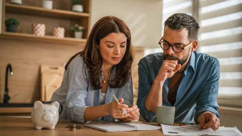 A couple reviews household finances together at a kitchen table, with paperwork, a notebook, coins and a piggy bank in front of them