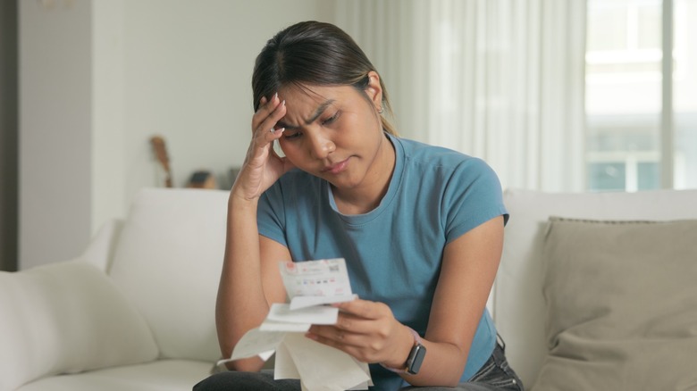 A concerned woman sits on a couch while looking at bills or receipts, with one hand pressed to her forehead