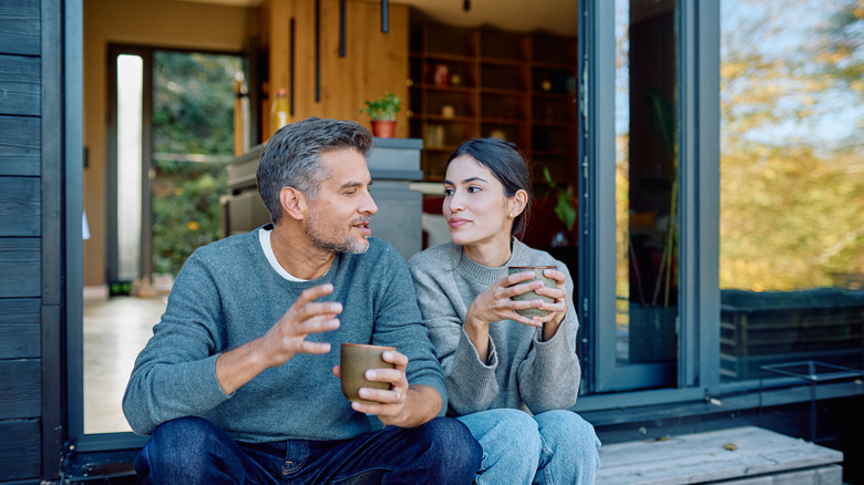 A couple sitting on an outdoor porch with mugs in their hands.