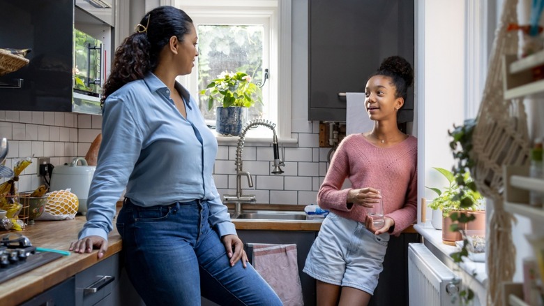 An adult and a child chatting in a bright kitchen, with the adult leaning on the counter and the child holding a glass near the window
