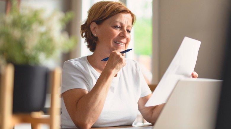 Middle-aged woman smiling while reviewing a document at a table, holding a pen with a laptop open in front of her