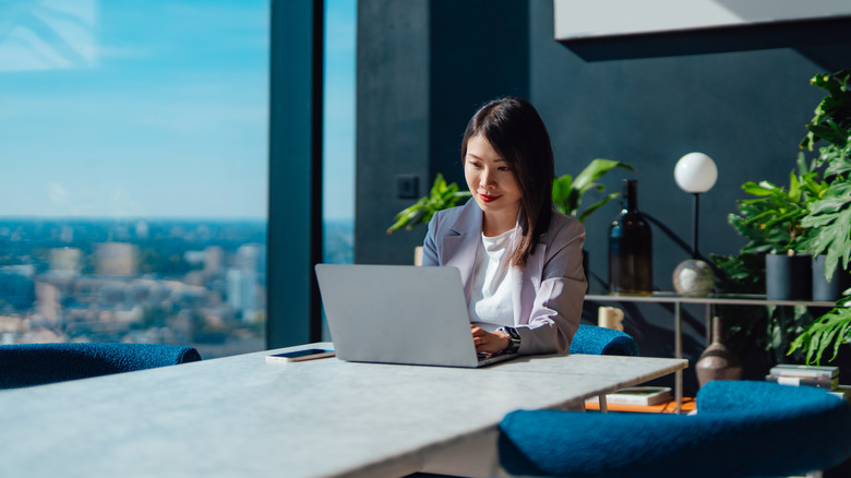 Young Asian businesswoman using laptop next to office window with cityscape.