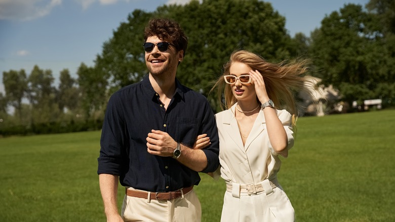 A beautiful young couple in elegant clothing stroll through a lush green field in a display of old-money style and rich people lifestyle.