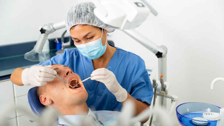 A masked dentist performing an oral examination as a man sits in a dentist's chair with his mouth open wide.