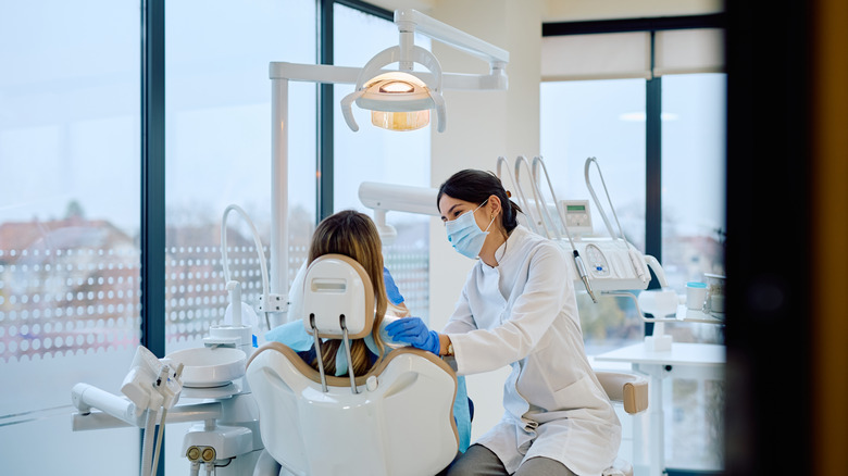 A masked dentist in a brightly lit dental office works on a patient sitting in a dentist's chair.