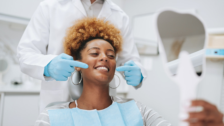 A smiling woman holding up a mirror in a dentist's chair while the dentist points at her teeth