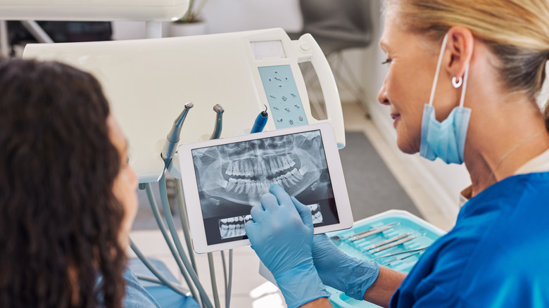 A dentist in blue scrubs examining a dental x-ray on digital tablet during a consultation.