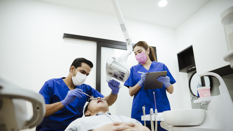 A man sits in a dentist's chair with his mouth open wide as a pair of dentists examine him.