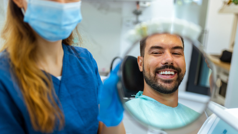 A masked dentist holding a mirror up to a smiling patient after a dental procedure.