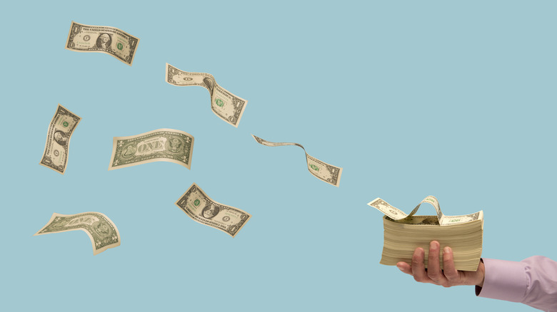 Man's hand holding stack of US currency with some bills flying away.
