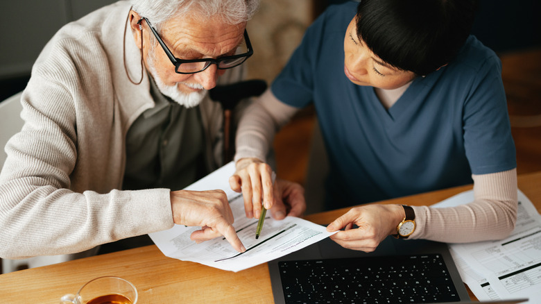 A caregiver explaining a document to an elderly man in front of a laptop.