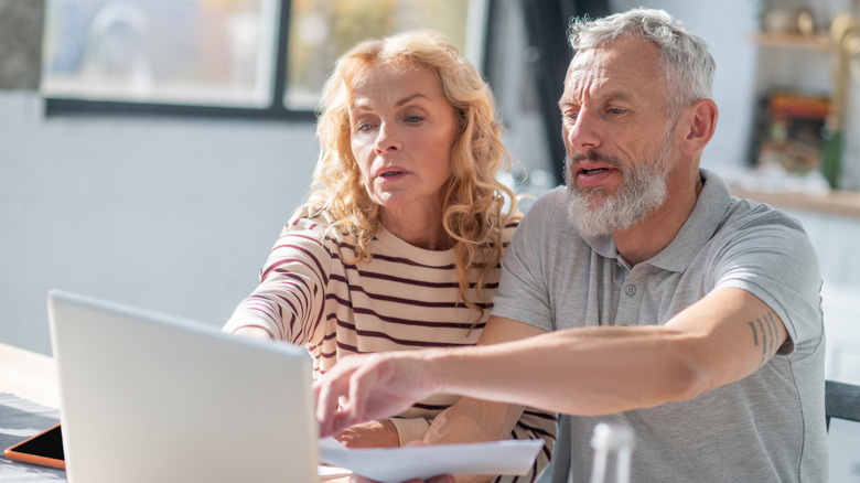 A mature couple looking at a laptop while holding documents.