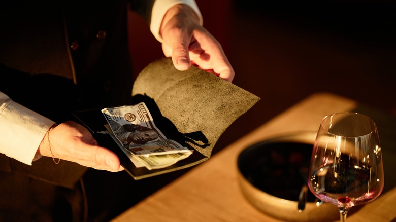 A server opens a bill holder showing cash beside a dining table with a wine glass and bowl