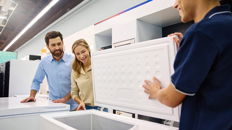 A couple is carefully examining a chest freezer while receiving advice from a sales associate